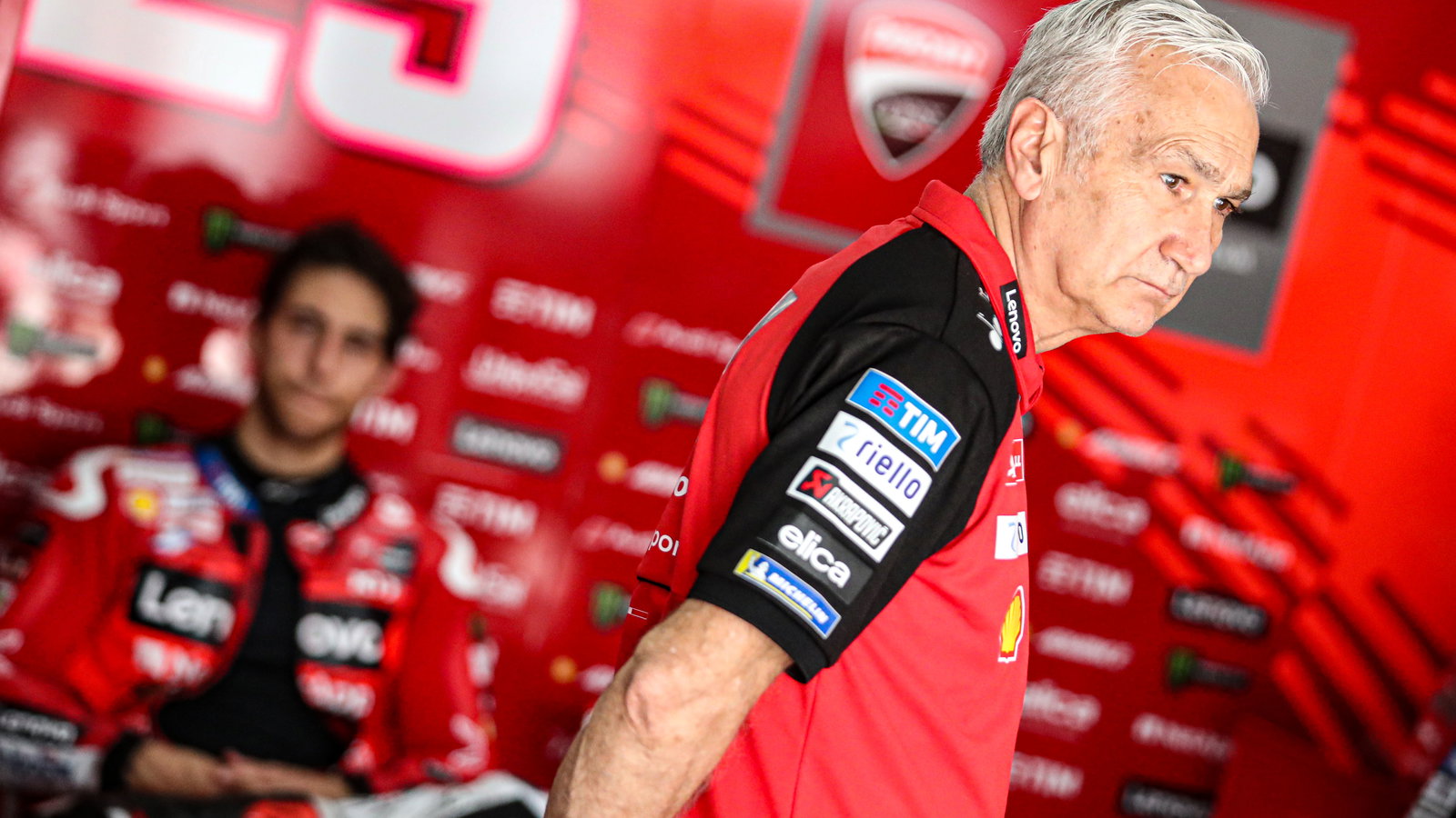 Davide Tardozzi stands in factory Ducati MotoGP garage with Enea Bastianini in the background. - Gold and Goose.